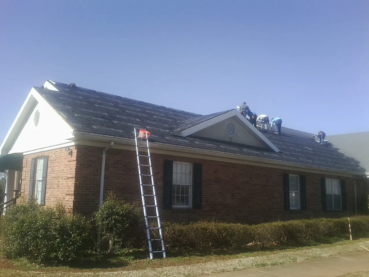 Crew tearing shingles off a brick ranch roof with a ladder set — Emergency Roof Repair work by our crew in Rainbow City