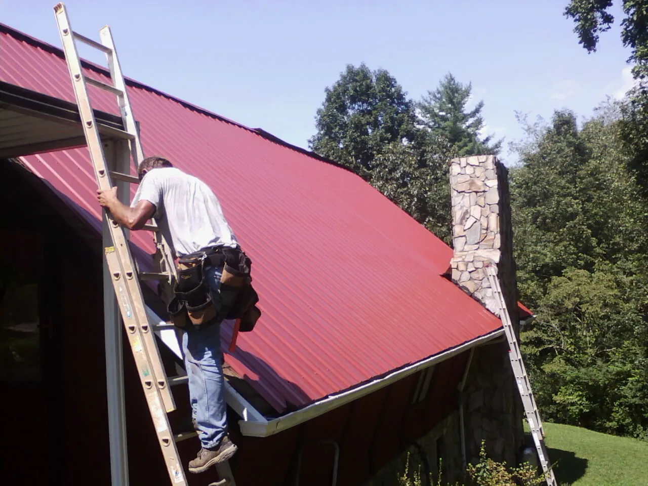 Roofer on a ladder beside a red metal roof and stone chimney — Soffit & Fascia Repair by Roofing Brothers in Rainbow City